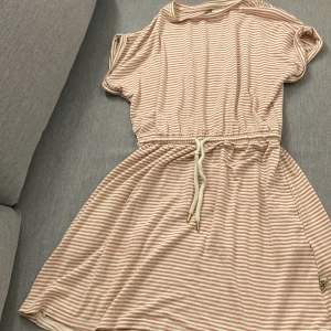 Randig klänning från Pomp de Lux - Säljer en söt randig klänning från Pomp de Lux i ljusrosa och vit med glittriga detaljer. Klänningen har korta ärmar och ett knytband i midjan för en bekväm passform. Perfekt för sommarens äventyr!