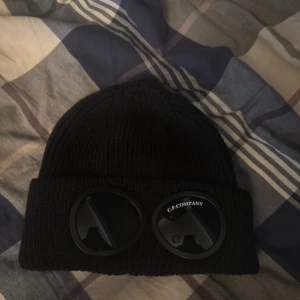 Cp mössa - Snygg svart mössa från C.P. Company med unika glasögondetaljer framtill. Tillverkad i mjuk bomull, perfekt för kyliga dagar. En stilren accessoar som sticker ut. Endast använd 2-3 gånger, köpt i local butiken i London för 1,5 år sedan.