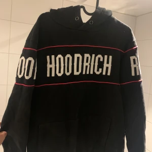 Svart hoodie från Hoodrich - Snygg svart hoodie från Hoodrich med vit text och röda detaljer. Den har en klassisk design med huva och långa ärmar. Perfekt för en avslappnad stil.