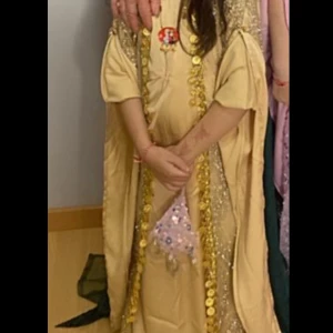 Guldfärgad klänning med glittriga detaljer - Elegant guldfärgad klänning med glittriga detaljer och korta ärmar. Klänningen har en unik design med guldfärgade dekorationer längs framsidan och ett knytband i ryggen.