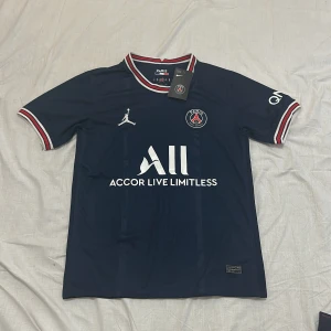 Mörkblå PSG Fotbollströja  - Snygg mörkblå PSG fotbollströja med korta ärmar och röd-vita detaljer på krage och ärmslut. Tröjan har Jordan-logga och PSG-emblem på bröstet samt texten 'Accor Live Limitless' på framsidan. Perfekt för fotbollsfans!