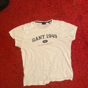 Vit t-shirt från Gant - Säljer en vit t-shirt från Gant med tryck 'GANT 1949' och 'NEW HAVEN CONNECTICUT' i svart och rött. T-shirten är kortärmad och har en klassisk rund halsringning.
