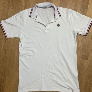 Moncler pikétröja  - Snygg vit pikétröja från Moncler med klassisk krage och röd-blå randiga detaljer. Tröjan har korta ärmar och en broderad logotyp på bröstet. Aldrig använt, står XXL men den är liten i storleken, den är mer för L