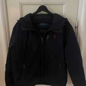 Svart dunjacka från Ralph Lauren - Snygg svart dunjacka från Ralph Lauren med broderad röd logotyp på bröstet. Jackan har dragkedja och är quiltad på insidan för extra värme. Perfekt för kyligare dagar.