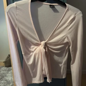 Ljusrosa knytblus från Gina Tricot - Säljer en söt ljusrosa blus från Gina Tricot med knytning framtill. Blusen har en v-ringad design och långa ärmar, perfekt för både vardag och festliga tillfällen.