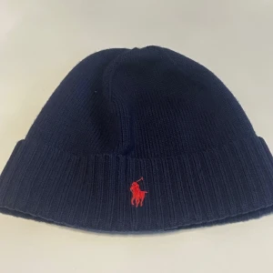 Mörkblå mössa från Polo Ralph Lauren - Snygg mörkblå mössa från Polo Ralph Lauren med röd broderad logotyp. Mössan är ribbad och perfekt för kyliga dagar.