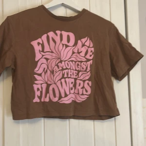 Brun croppad t-shirt från H&M - Säljer en brun croppad t-shirt från H&M med texten 'Find Me Amongst The Flowers' i rosa. T-shirten har korta ärmar och en avslappnad passform. Perfekt för en avslappnad stil.