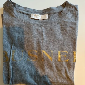 Grå t-shirt från Busnel - Snygg grå t-shirt från Busnel med guldtryck på framsidan. Perfekt för en avslappnad stil med en touch av lyx. Gjord i mjukt material för extra komfort.
