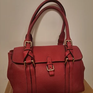 Röd handväska med spänne - Snygg röd handväska i skinn med dubbla handtag och dekorativa spännen. Väskan har en stilren design med metallfötter på undersidan för extra skydd. Perfekt för att ge en färgklick till din outfit.