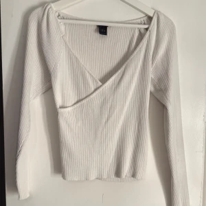 Vit ribbad omlottopp - Säljer en stilren vit ribbad omlottopp med långa ärmar. Perfekt för en chic och enkel look. Toppen har en omlottdesign framtill som ger en fin detalj.