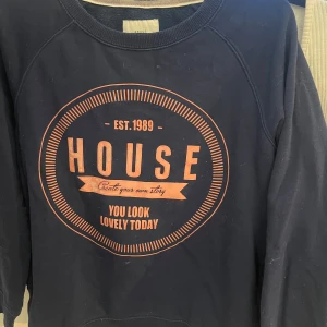 Mörkblå sweatshirt med tryck - Säljer en mörkblå sweatshirt med ett stort tryck i orange på framsidan. Texten 'HOUSE' och 'You look lovely today' ger en unik touch. Tröjan har långa ärmar och en rund halsringning. Perfekt för en avslappnad stil.