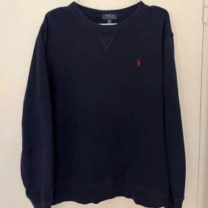 Mörkblå tröja från Polo Ralph Lauren - Säljer en stilren mörkblå tröja från Polo Ralph Lauren med det klassiska röda logotypbroderiet på bröstet. Tröjan har långa ärmar och en rund halsringning. Perfekt för en avslappnad och snygg look.