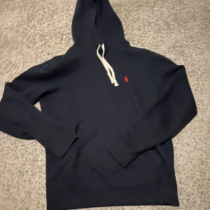 Mörkblå hoodie från Ralph Lauren - Snygg mörkblå hoodie från Ralph Lauren med en liten röd logga på bröstet. Den har en justerbar huva med vita snören och långa ärmar. Perfekt för en avslappnad stil.