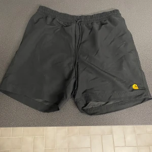 Svarta badshorts från Carhartt - Snygga svarta badshorts från Carhartt med gul broderad logga. De har en elastisk midja med snörning och en bakficka. Perfekta för sommarens badäventyr!