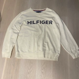 Vit tröja från Tommy Hilfiger - Säljer en stilren vit tröja från Tommy Hilfiger med mörkblå text på framsidan. Tröjan har långa ärmar och en klassisk rund halsringning med ribbade muddar. Perfekt för en avslappnad look.