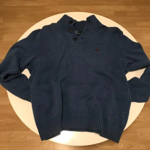 Blå tröja från Polo Ralph Lauren - Snygg blå tröja från Polo Ralph Lauren med ribbad krage och knappar. Den har långa ärmar och ett diskret rött broderat märke på bröstet. Perfekt för kyligare dagar.