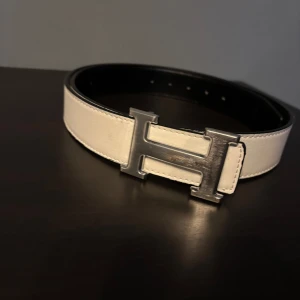 Vitt skärp med H-spänne - Snyggt vitt skärp med ett stort H-format spänne i silver. Perfekt för att ge en stilren touch till din outfit. Skärpet är gjort av skinn och har en elegant design.