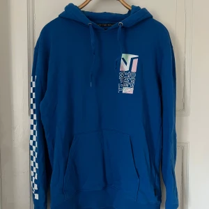 Blå hoodie från Vans - Säljer en blå hoodie från Vans med texten 'Off The Wall' på bröstet och ryggen. Den har en klassisk känguruficka och justerbar huva. Ärmen har ett schackrutigt mönster som ger en cool touch. Perfekt för en avslappnad stil.
