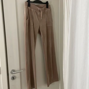 Beige mjukisbyxor från Juicy Couture - Säljer ett par snygga beige mjukisbyxor från Juicy Couture. Byxorna har en bekväm passform med dragsko i midjan och fickor både fram och bak. Perfekta för en avslappnad stil.