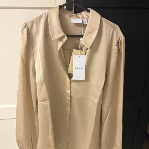 Beige satinblus från Vila - Elegant beige satinblus från Vila med långa ärmar och klassisk krage. Perfekt för en stilren look.