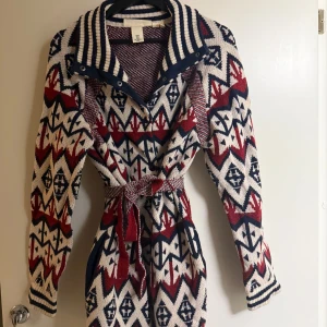 Mönstrad stickad kappa från H&M - Säljer en snygg stickad kappa från H&M med ett färgglatt mönster i rött, blått och vitt. Den har långa ärmar och ett knytbälte i midjan för en fin passform. Perfekt för att hålla sig varm och stilren under kyliga dagar.