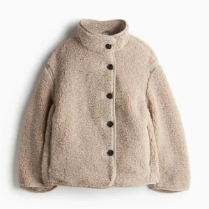 Beige teddyjacka med knappar - Mysig beige teddyjacka med knappar framtill. Perfekt för kyliga dagar med sin fluffiga och varma design. Jackan har en hög krage och långa ärmar för extra värme.