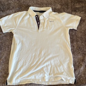 Vit kortärmad pikétröja från GANT - Säljer en klassisk vit pikétröja från GANT med korta ärmar och krage. Tröjan har en knappslå med mörka detaljer och GANT-loggan broderad på bröstet. Perfekt för en stilren look. Den har ett litet håll och det är den på bilden men annars är det helt oskad.