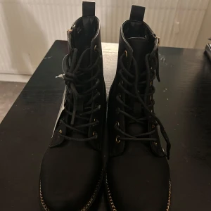 Svarta boots med dragkedja och snörning - Snygga svarta boots med både snörning och dragkedja för enkel på- och avtagning. De har en robust sula och gulddetaljer som ger en stilren look. Perfekta för höst och vinter.