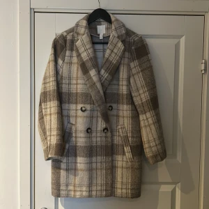 Rutig kappa från H&M - Säljer en stilren rutig kappa från H&M i beige och bruna toner. Den har dubbelknäppning med knappar och två fickor framtill. Perfekt för höst och vår. 