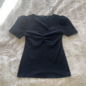 Svart ribbad topp med puffärmar - Säljer en snygg svart ribbad topp med korta puffärmar och en v-ringad design. Toppen har en smickrande passform och är perfekt för alla tillfällen. Den är i bra skick och knappt använd. 