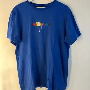 En blå t-shirt med en färgglad droppande logotyp av märket Ellesse. T-shirten har en rund halsringning och korta ärmar. T-shirten är lite lätt blekt i sömmen upp till, se bild 5. Annars hel och ren