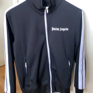 Svart jacka från Palm Angels - Säljer en snygg svart jacka från Palm Angels med vita ränder längs ärmarna. Jackan har en dragkedja framtill och är långärmad. Perfekt för en casual look eller träning. Den är i nyskick och redo för en ny ägare!