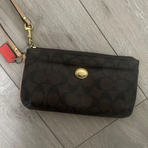 Brun clutch från Coach - Säljer en snygg brun Clutch från Coach med deras klassiska mönster. Den har en praktisk dragkedja och flera fack inuti för kort och kontanter. Perfekt för både vardag och fest! 👜