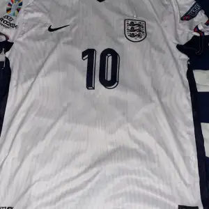 Säljer en vit England fotbollströja från Nike med nummer 10 på framsidan och Bellingham på baksidan. Tröjan har korta ärmar och är i nyskick. Perfekt för fotbollsfans som vill visa sitt stöd!