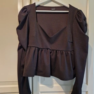 Svart topp från Gina Tricot - Säljer en snygg svart topp från Gina Tricot i storlek XL. Toppen har en v-ringad design med långa ärmar och en volangdetalj i midjan. Perfekt för både vardag och fest!