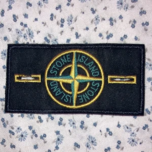 Svart broderad patch från Stone Island - Säljer en snygg svart broderad patch från Stone Island. Den har det klassiska kompassmönstret i gult och grönt. Perfekt för att sätta på jackor, byxor, väskor och med mera.