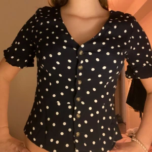 Svart prickig blus med knappar - Snygg svart blus med vita prickar och knappar framtill. Den har korta ärmar och en v-ringad design som ger en klassisk look. Perfekt för en stilren outfit.