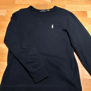 Blå tröja från Polo Ralph Lauren - Säljer en stilren mörkblå tröja från Polo Ralph Lauren med det klassiska logotypbroderiet på bröstet. Tröjan har långa ärmar och en rund halsringning med dekorativ söm. Perfekt för en avslappnad och snygg look.