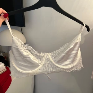 Vit siden bralette med spetsdetaljer - Elegant vit bralette i siden med vackra spetsdetaljer längs kanterna. Den har justerbara axelband och en mjuk, glansig yta som ger en lyxig känsla. Perfekt för att lägga till en touch av elegans under dina kläder.