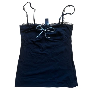 Svart linne med spetsdetaljer - Snyggt svart linne med smala axelband och spetsdetaljer längs kanten. Linnet har en söt rosett framtill som ger en extra touch. Perfekt för en stilren look. Passar en XS eller S bäst! 💓