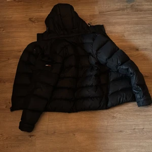 Marinblå  dunjacka från Tommy Hilfiger - Säljer en marinblå dunjacka från Tommy Hilfiger med huva och dragkedja. Jackan har en klassisk märkeslogga på bröstet men även på vänstra armen. Perfekt för kyliga dagar. Luvan är avtagbar och jackan har inga synliga difekter eller andra difekter.