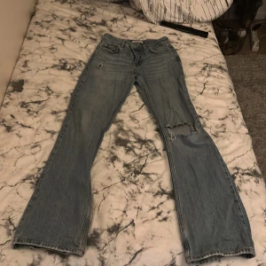 Blå jeans med slitningar low waist boot cut  - Säljer ett par blå jeans från Perfect Jeans med slitningar på ena benet. De har en klassisk femficksdesign och en bekväm passform. Perfekta för en avslappnad stil.