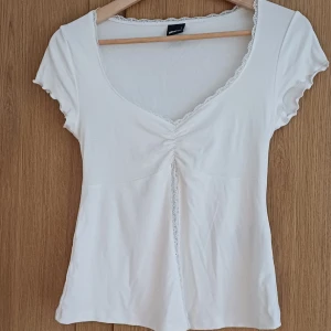 Vit topp med spetsdetaljer - Säljer en söt vit topp från ginatricot med spetsdetaljer längs v-ringningen och framtill. Toppen har korta ärmar. Perfekt för en somrig look!