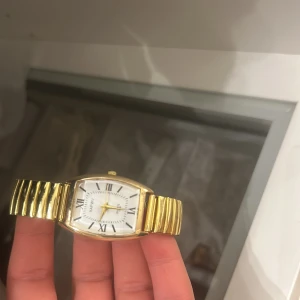 Guldfärgad klocka med vit urtavla - Elegant guldfärgad klocka med en vit urtavla och romerska siffror. Klockan har ett flexibelt armband i metall och en klassisk design som passar perfekt för en stilren look.