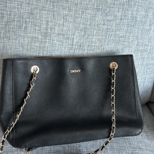 Svart axelväska från DKNY - Snygg svart axelväska från DKNY med kedjedetaljer och dragkedja. Perfekt för att bära dina dagliga nödvändigheter med stil. Väskan har en elegant design och är tillverkad i skinn.