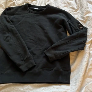 Svart sweatshirt från CP Company - Snygg svart sweatshirt från CP Company med rund hals och en unik detalj på ärmen. Perfekt för en stilren och avslappnad look. Storlek 158