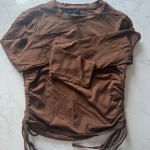 Brun långärmad topp med knyt - Snygg brun långärmad topp från PIECES med knyt i sidorna för en justerbar passform. Perfekt för en avslappnad stil med en twist. Passar bra till jeans eller kjol.