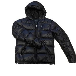 Peak Performance jacka  - Snygg och varm svart dunjacka från Peak Performance med huva och dragkedja. Perfekt för kalla dagar med sin pufferdesign och praktiska fickor med dragkedjor. Hör av dig vid mer frågor!