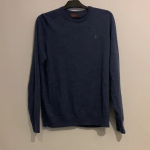 Blå tröja i merinoull från Morris - Säljer en stilren blå tröja från Morris i 100% merinoull. Tröjan har en liten broderad detalj på bröstet och långa ärmar. Perfekt för kyligare dagar.