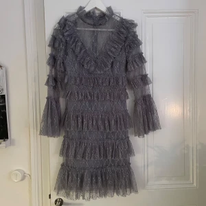 By malina blå klänning  - Elegant blå klänning i spets med volanger och långärmad design. Den har en v-ringad hals och flera lager av volanger som ger en romantisk känsla. Perfekt för speciella tillfällen. Helt oanvänd med prislapp kvar. Ordinarie pris 2800kr men slutsåld.